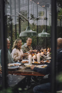 Retired male and female friends enjoying dinner party together seen through glass
