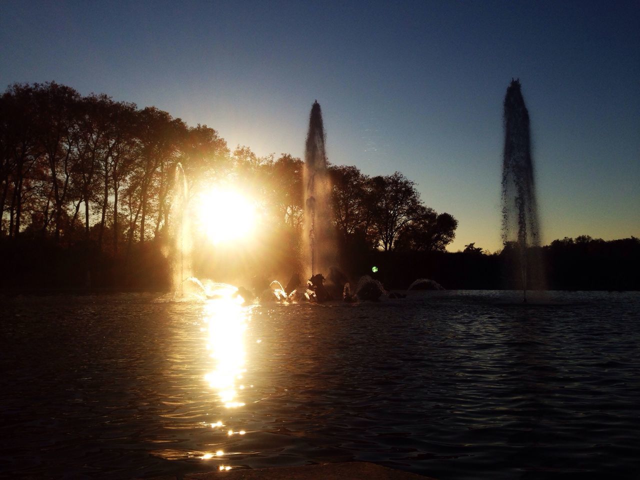 sun, water, waterfront, architecture, building exterior, built structure, clear sky, reflection, city, lens flare, sunlight, silhouette, sunset, tree, sunbeam, skyscraper, tall - high, river, tower, travel destinations