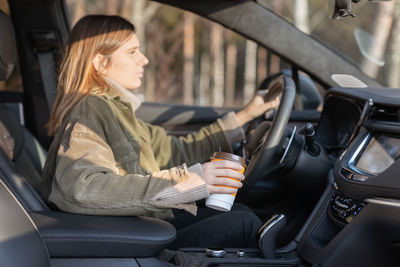 Cute young girl driving car by one hand and drinking coffee. woman neglects safety in the vehicle