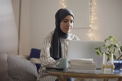 Young businesswoman wearing hijab while working on laptop at table in living room