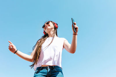 From below latin american female in white t shirt and modern sunglasses with cellphone and wireless headphones on blue background on sunny day