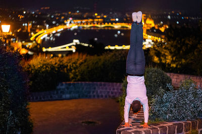 Rear view of man doing handstand on retaining wall against illuminated city at night