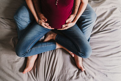 Low section of pregnant couple sitting on bed at home