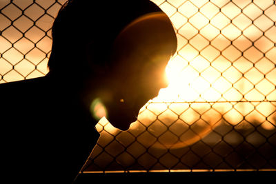 Silhouette man seen through chainlink fence against sky during sunset