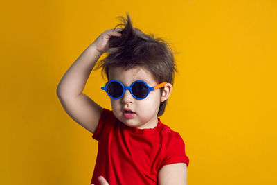 Cheerful baby boy in red t-shirt stands on yellow background in sunglasses