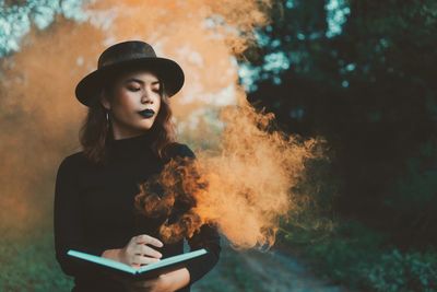 Young woman holding book while standing against trees in park