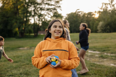 Smiling girl holding blue and yellow balls while standing in playground at summer camp