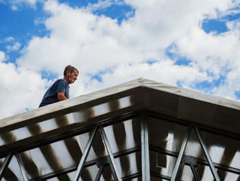 Low angle view of man standing on roof against cloudy sky