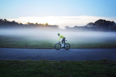 Side view of man cycling on street amidst grassy field in foggy weather