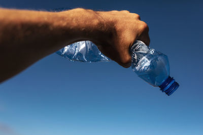Cropped hand of person holding crumpled bottle against clear blue sky