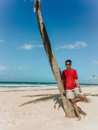 Full length portrait of young man leaning on palm tree trunk at beach against blue sky
