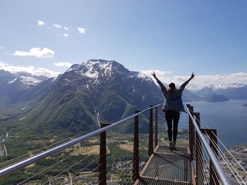 Rear view of woman with arms raised standing on footbridge against mountains