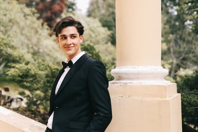 A beautiful young man, the groom in an elegant wedding suit, stands posing in the city's old park