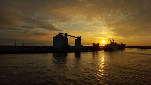 Silhouette factory by sea against sky during sunset