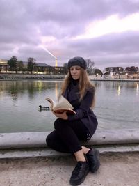 Young woman sitting by lake against sky