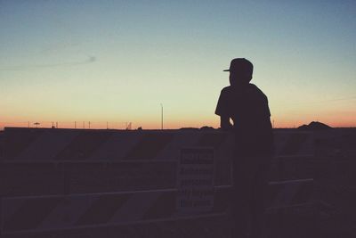 Rear view of silhouette man standing by barricade on field against sky during sunset