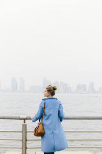 Young woman looking at view while standing by railing and river in city against clear sky