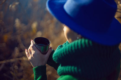 Woman in knitted sweater and blue hat stands bushes sunset and drinks tea from an iron thermos mug