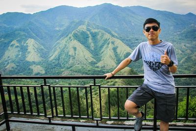 Portrait of young man showing thumbs up while standing against mountain