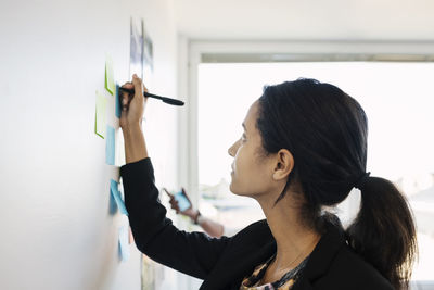 Businesswoman writing in adhesive note on wall in office