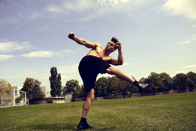 Shirtless man kicking on field against sky