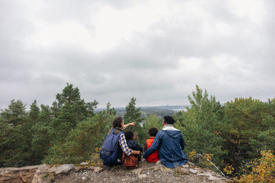 Rear view of woman showing trees to family while sitting on cliff against sky