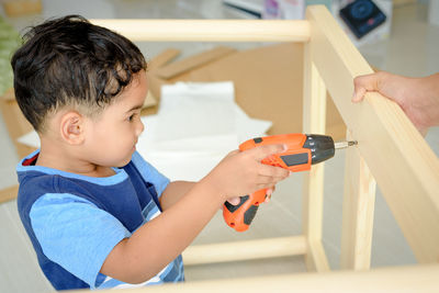 Boy drilling wood held by person at home