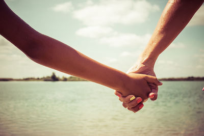 Cropped image of mother and child holding hands by lake against sky