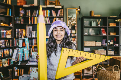 Portrait of smiling girl holding toy pencil and set square while standing in bookstore