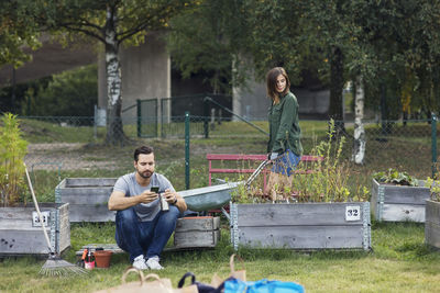 Mid adult man using mobile phone while woman working in urban garden