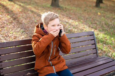 An emotional boy talks on the phone covers his mouth with his hand and laughs sitting on a park 