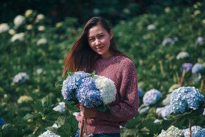 Portrait of smiling woman holding flowers while standing by plants