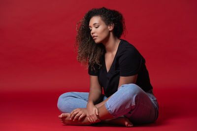 Full body of young overweight african american woman in casual black shirt and jeans with bare feet sitting in red studio looking down