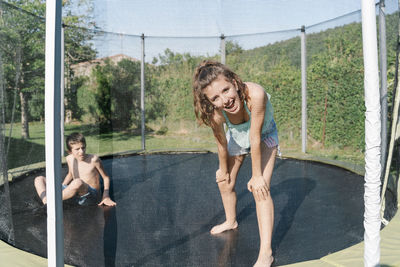 Horizontal photo of a blonde boy and a brunette girl looking at camera having fun on a trampoline on a green background. the teenager and the child wear short jeans and they look happy