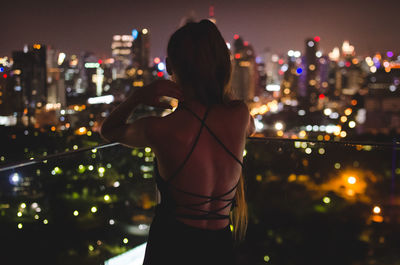 Rear view of woman looking at illuminated cityscape while standing on balcony