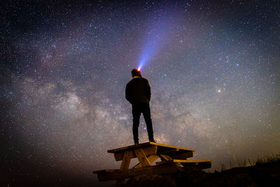 Silhouette of man stargazing with headlamp standing on a picnic table.