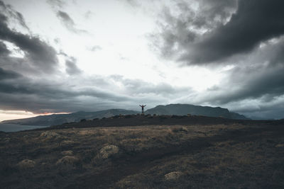 Mid distance view of man with arms outstretched standing on landscape against cloudy sky