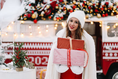 A beautiful woman with red lips holds gift boxes at a decorated christmas van on the street in city