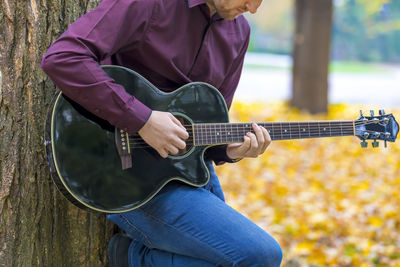 Midsection of young man playing guitar while standing by tree in park during autumn