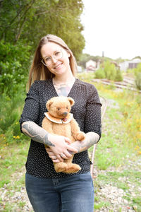 A young caucasian girl with glasses and tattoos smiles, a brown teddy bear