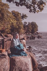 Woman sitting on rock at shore against sky