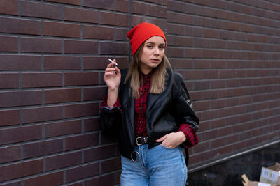 Portrait of young woman wearing knit hat smoking cigarette while standing against brick wall