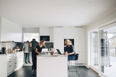 Woman and teenage girl using laptops while man boy standing in kitchen at home