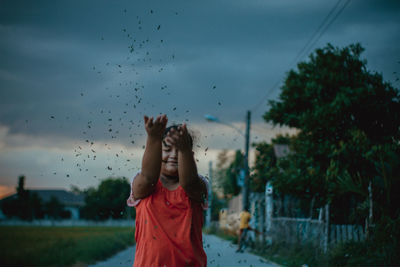 Girl throwing leaves while standing on road against cloudy sky at dusk