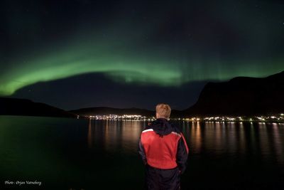 Rear view of man standing by illuminated lake against sky at night