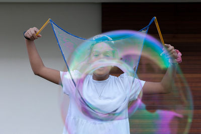 Cheerful young african american female teenager with dark hair in white t shirt playing with big colorful soap bubbles and having fun standing on street near wooden wall