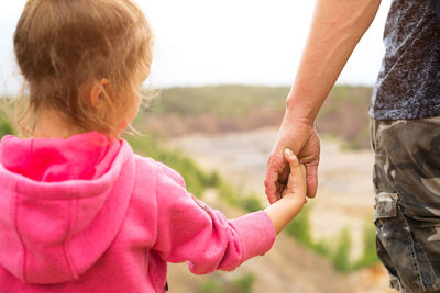 Daughter in a pink hoodie holding dad's hand, standing on top of a mountain with a beautiful 