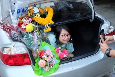 Cropped hands photographing young woman with bouquet in car trunk
