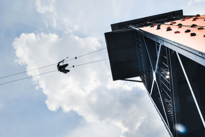 Low angle view of man hanging on rope by buildings against cloudy sky