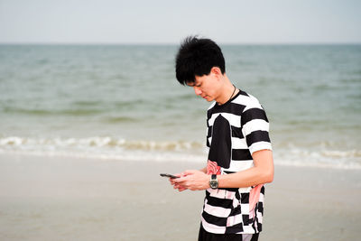 Portrait of young man stands on the beach and uses the mobile phone with blurred seascape background